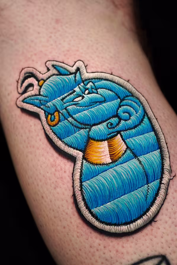 Tattoo on an arm designed to look like an embroidered patch, depicting the Genie from Aladdin in blue with gold accents, styled in a cartoon embroidery effect