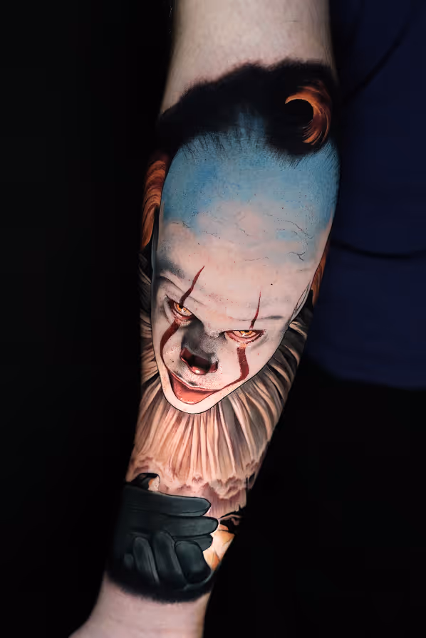 Realistic forearm tattoo of Pennywise the clown with orange eyes, red makeup, and a ruffled collar reaching out a gloved hand.